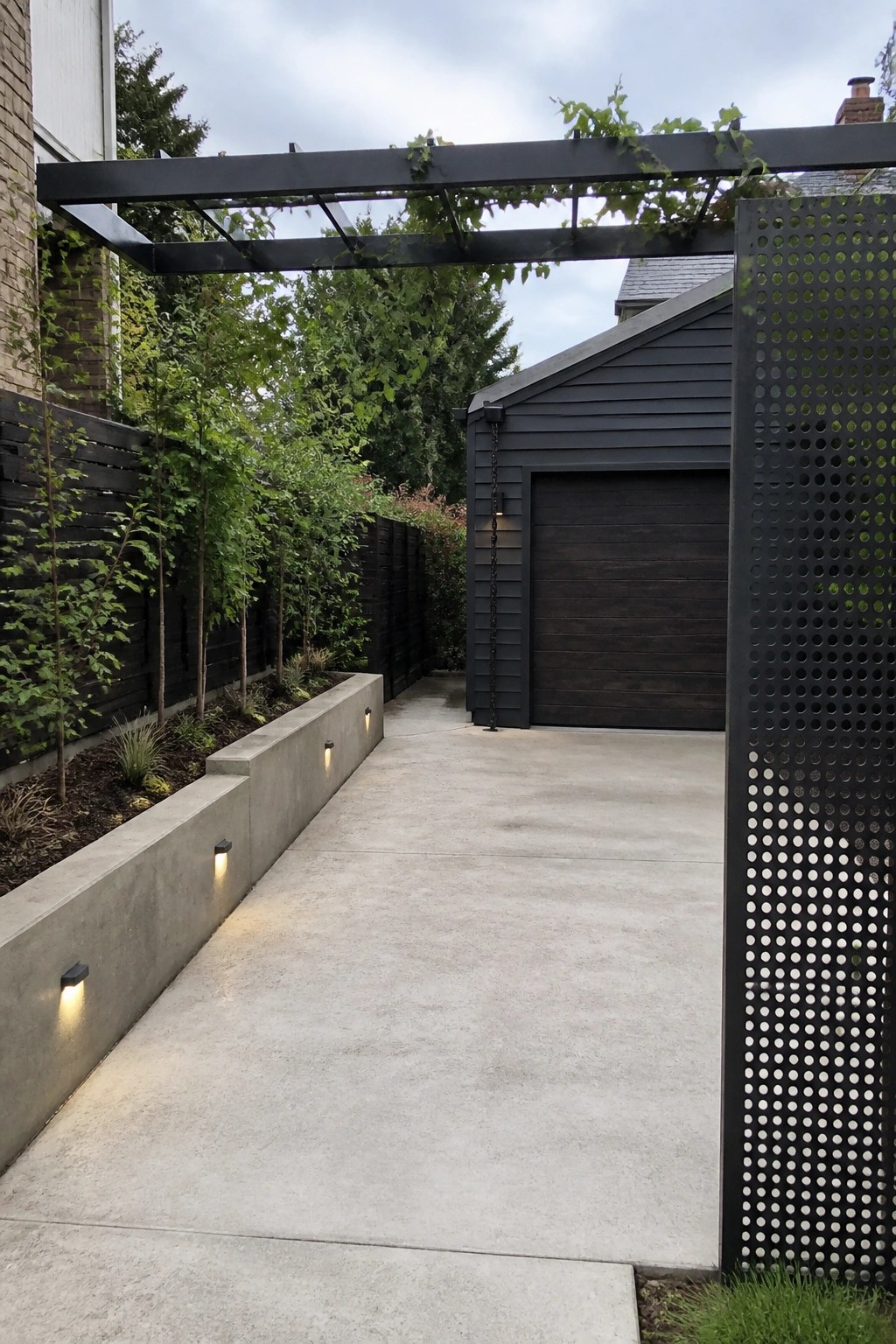 A concrete driveway path runs between a low concrete wall with recessed lights on the left and a tall perforated metal screen on the right, leading to a dark wood garage under a vine-covered pergola.