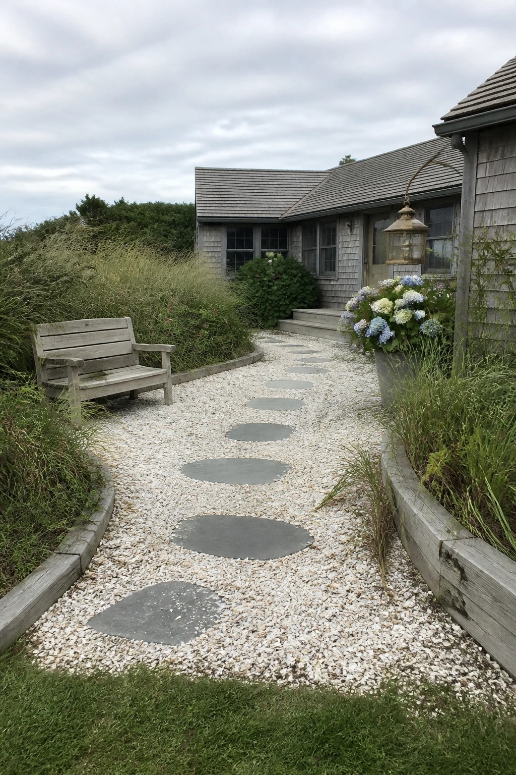 A gravel path with large flat stepping stones leads to a gray shingled house, bordered by wooden edging, a bench on the left, and potted hydrangeas on the right.