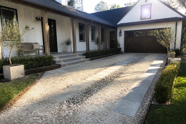A gravel driveway with parallel strips of large gray stone pavers leading toward a white house with a wooden garage and covered porch.