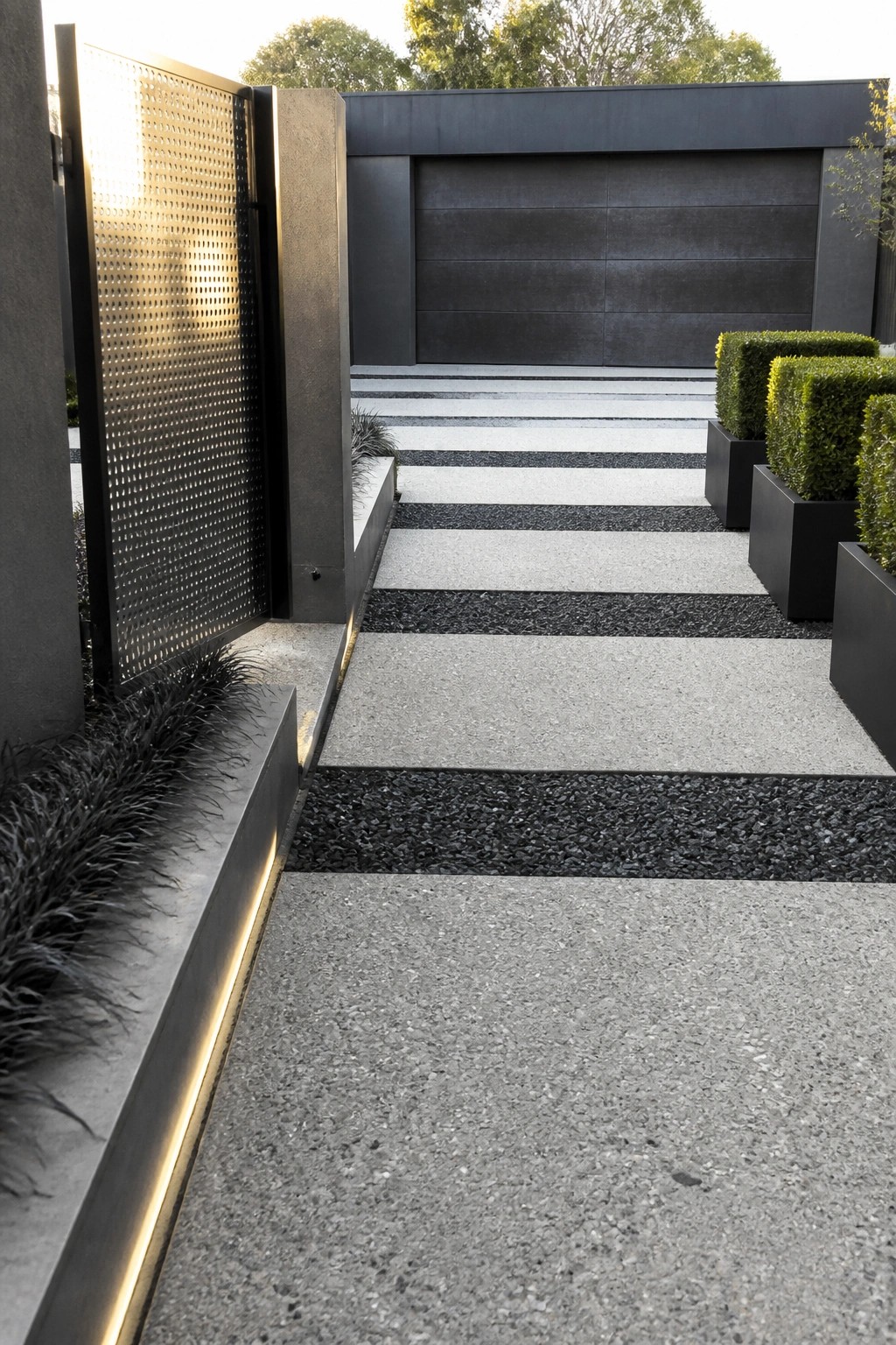 A modern exterior pathway made of alternating light concrete slabs and dark gravel strips, bordered by metal planters with shrubs and a perforated metal gate on the left.