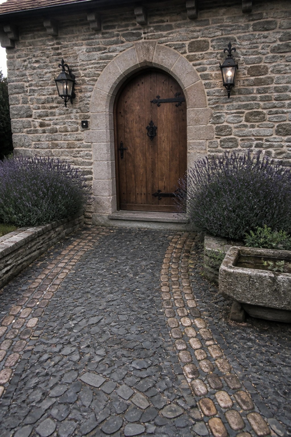 A cobblestone path made of mixed irregular stones leads to an arched wooden door on a stone house, flanked by lavender bushes and two lanterns.
