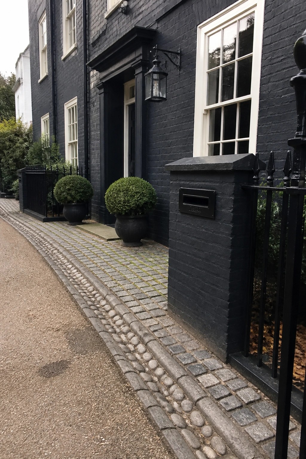 Black brick house with white windows, potted shrubs, lantern, mailbox, and cobblestone path