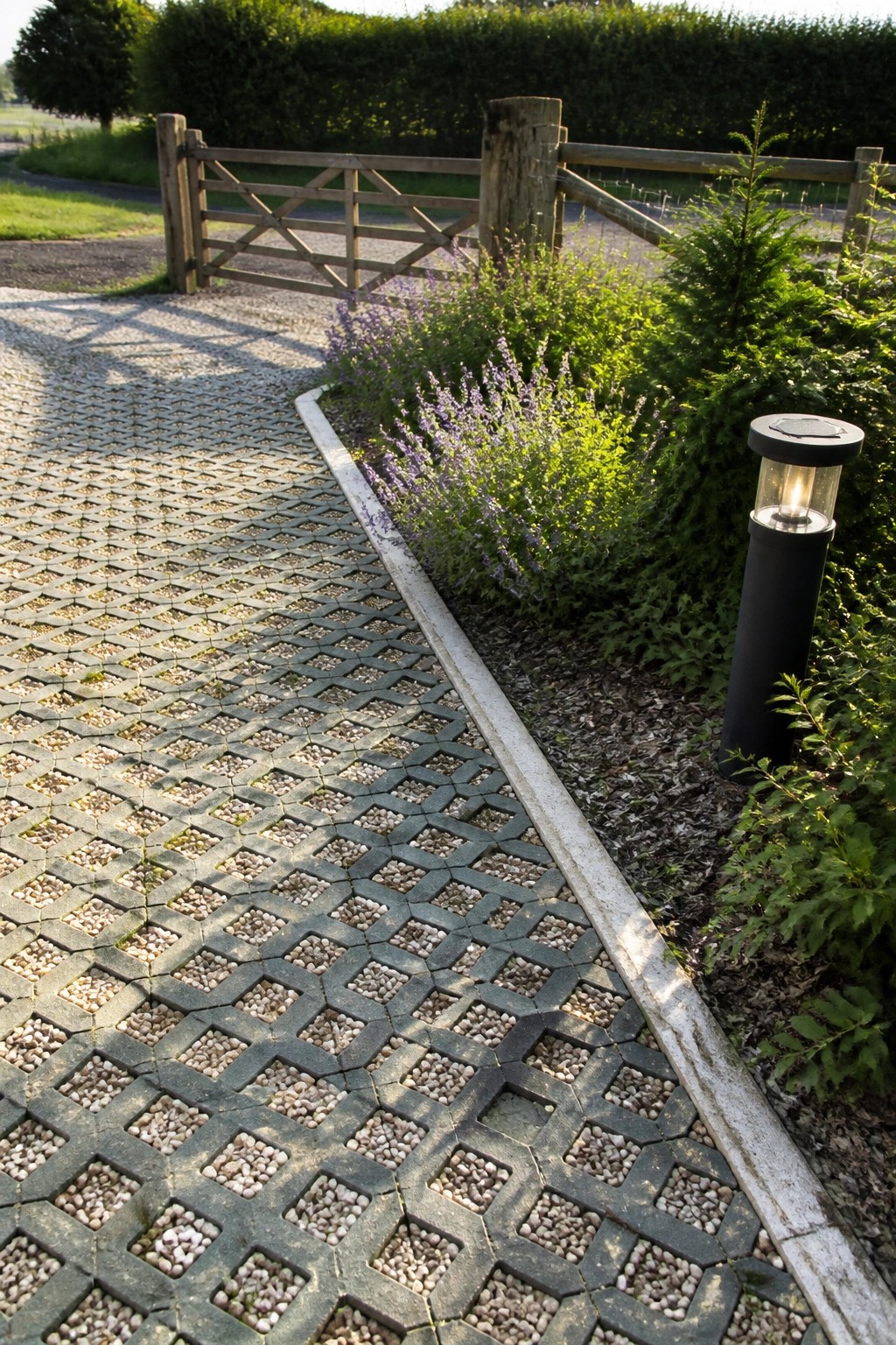 A gravel-filled grid paver path runs alongside a garden bed with purple flowers and leads toward a wooden gate.