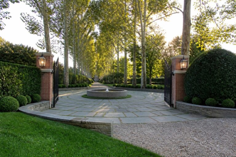19 Elegant Driveway Entrance Ideas That Make a Grand First Impression A wide stone driveway passes through open black metal gates set in brick pillars, with a round stone fountain centered in the foreground and a long double row of trees leading toward a house in the distance.