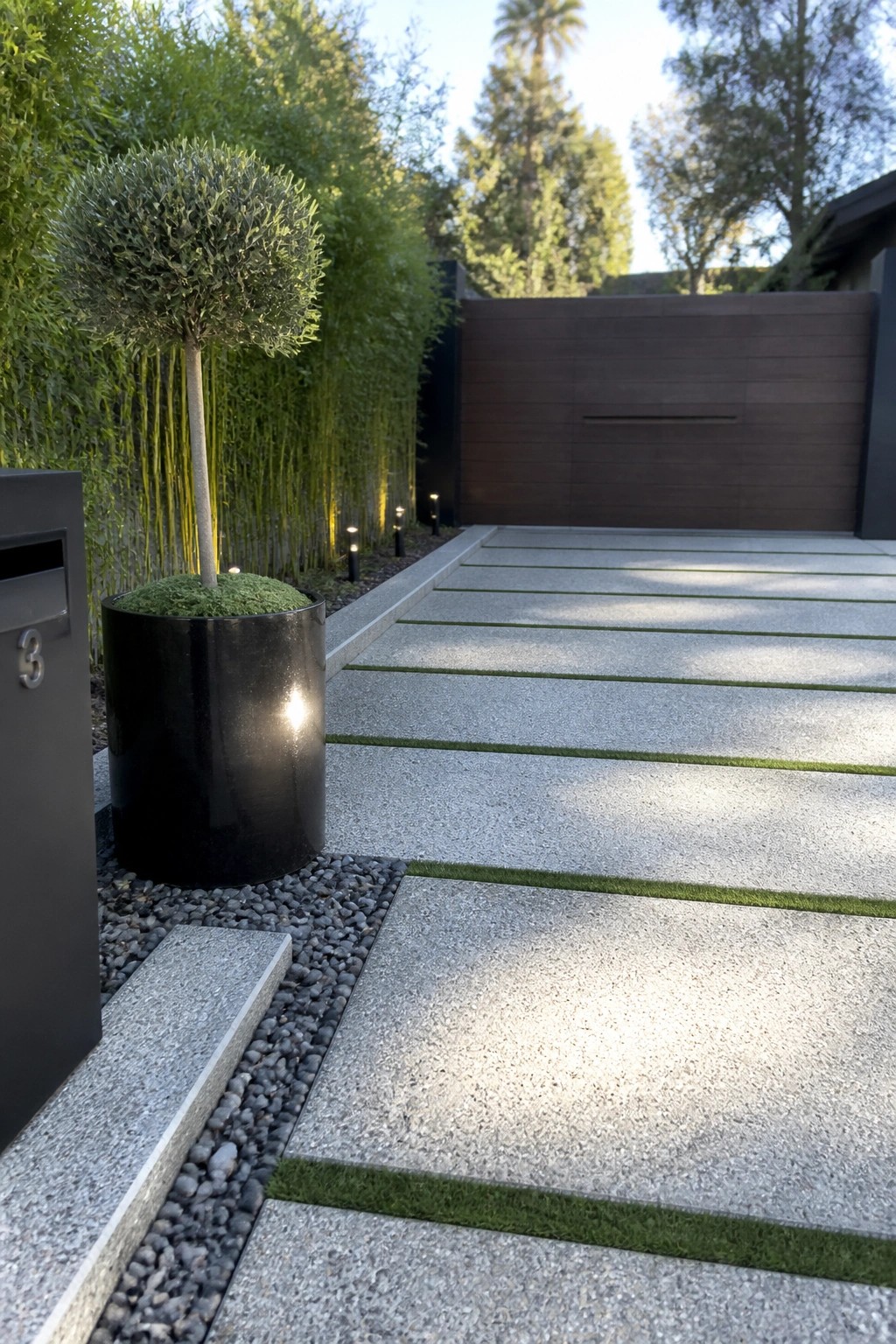 A driveway made of large gray concrete pavers with narrow grass strips between them leads to a dark wooden gate, with a black planter holding a small tree on the left side.