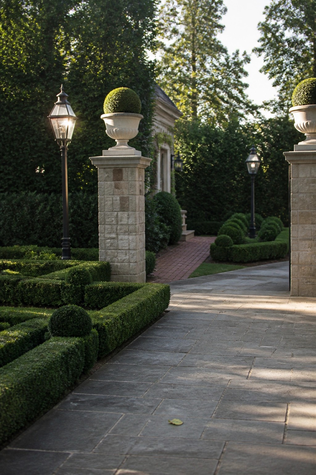 Paved walkway through formal garden with hedges, topiary urns, and glowing lanterns.