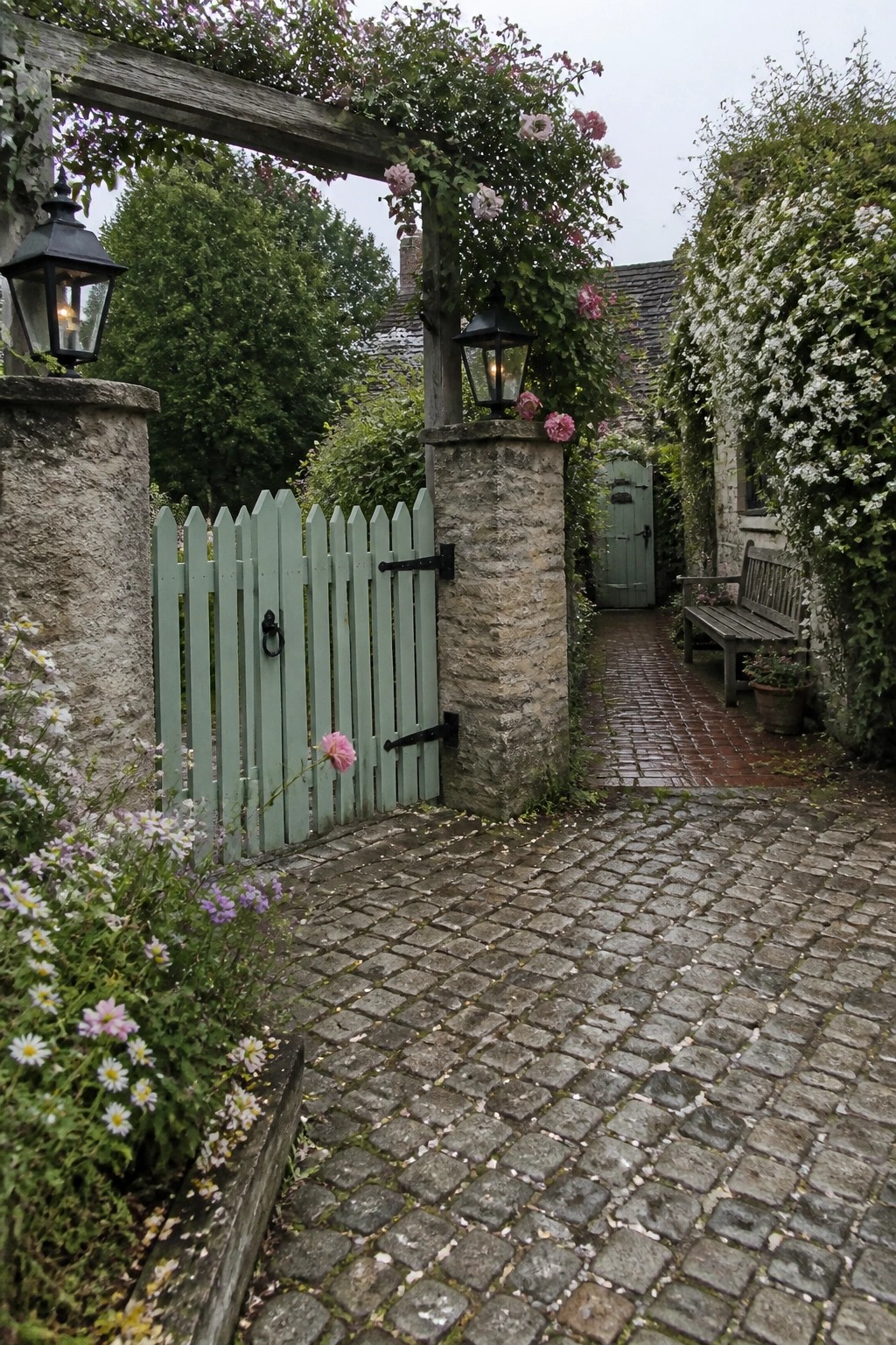A light green wooden picket gate set between rough stone pillars on a cobblestone driveway path, with black lanterns on the pillars and pink climbing roses overhead.