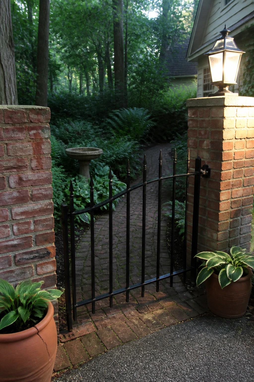 Black iron gate between brick pillars with lantern, potted plants, and garden path