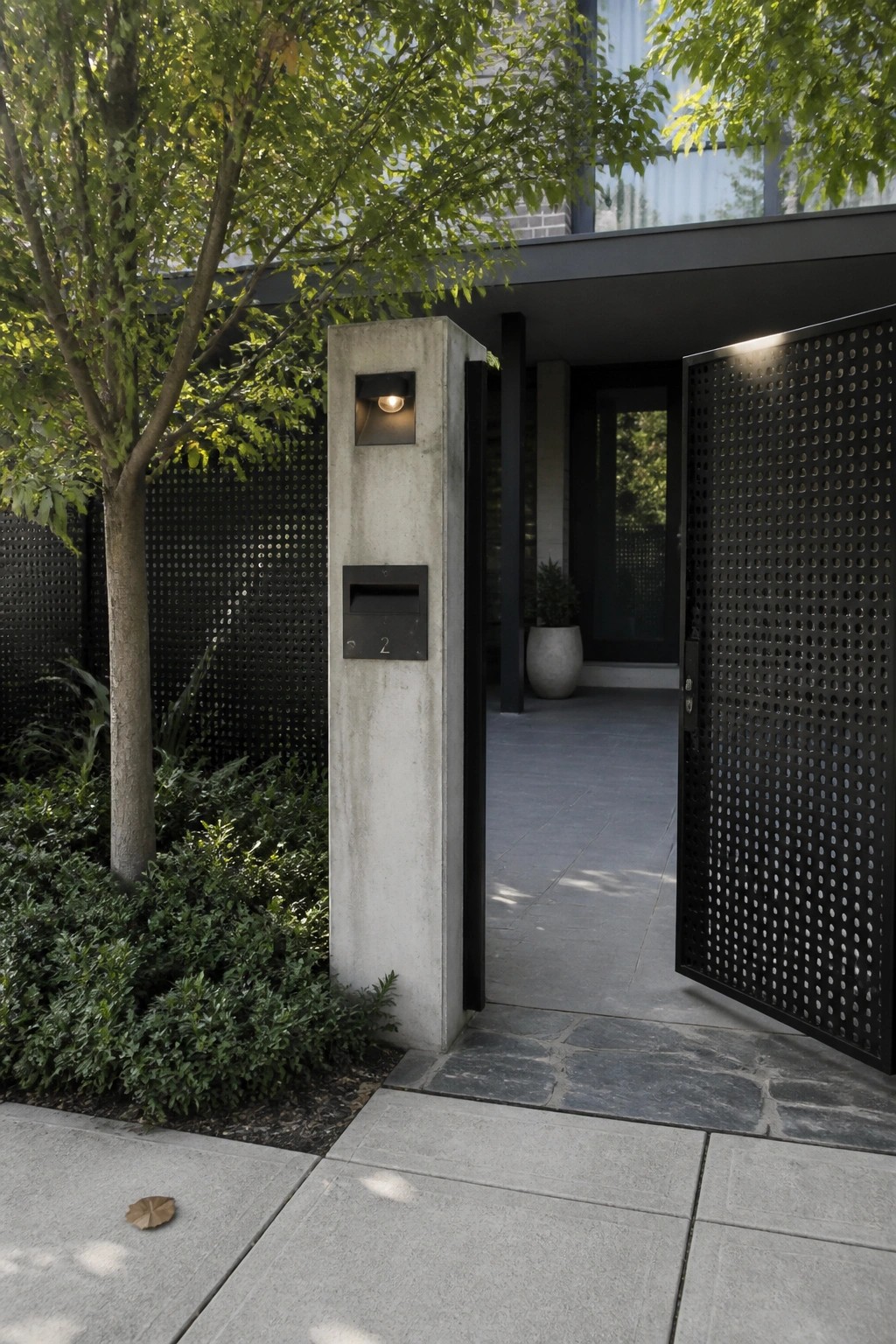 A black perforated metal gate stands open next to a tall concrete pillar that holds an integrated mailbox and light, with stone paving and greenery around the entrance.