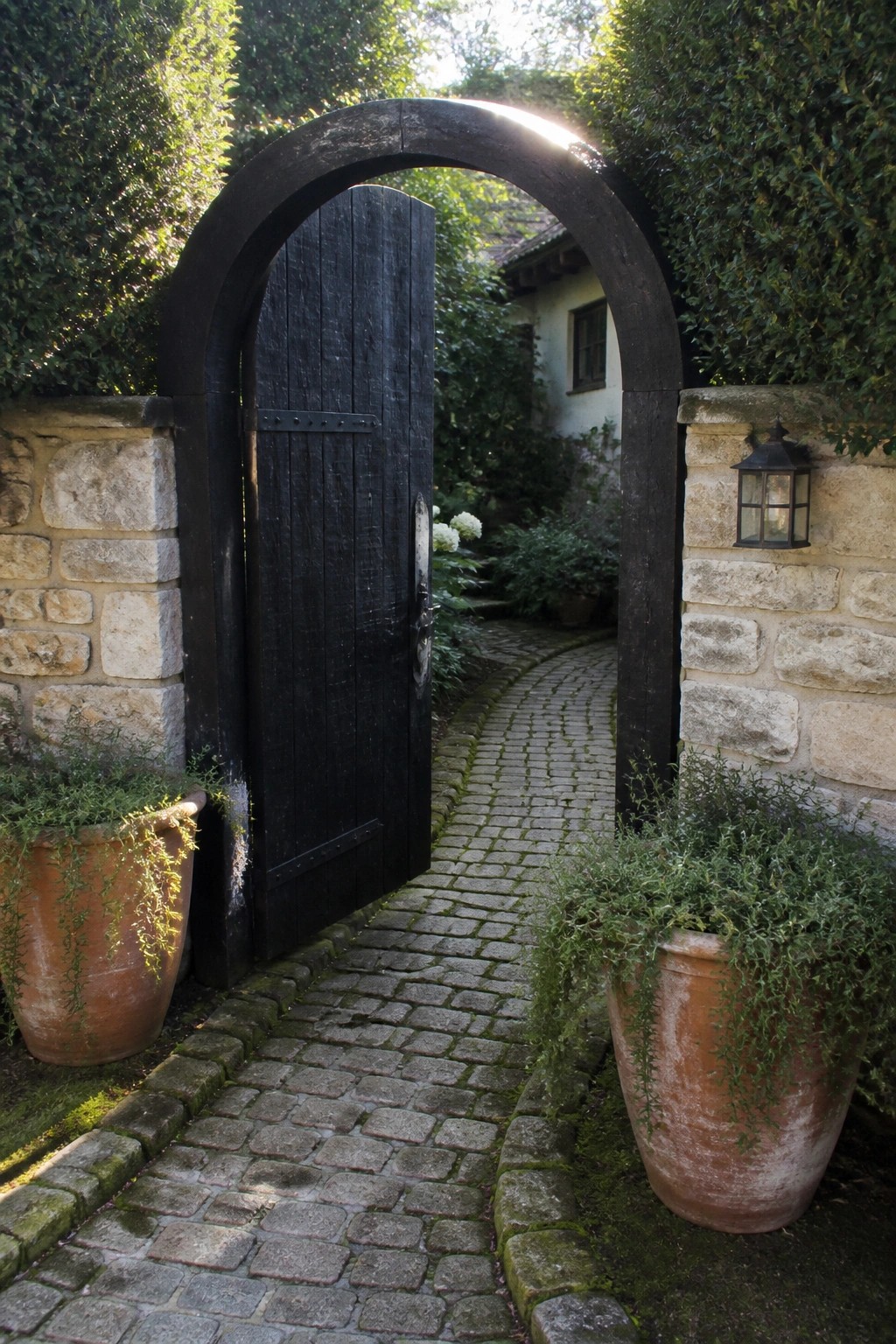 A black wooden gate inside a stone archway opens onto a cobblestone path with terracotta pots on either side.