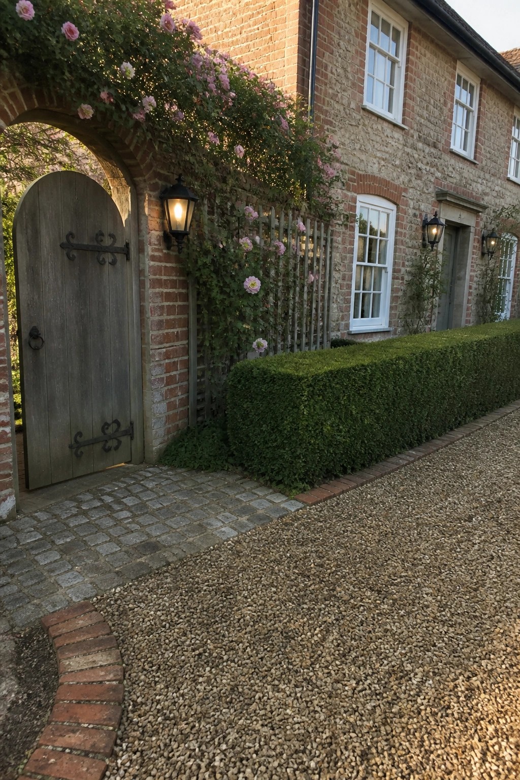 A pea gravel area bordered by a clipped green hedge sits in front of a brick house with an arched wooden gate and climbing pink roses.