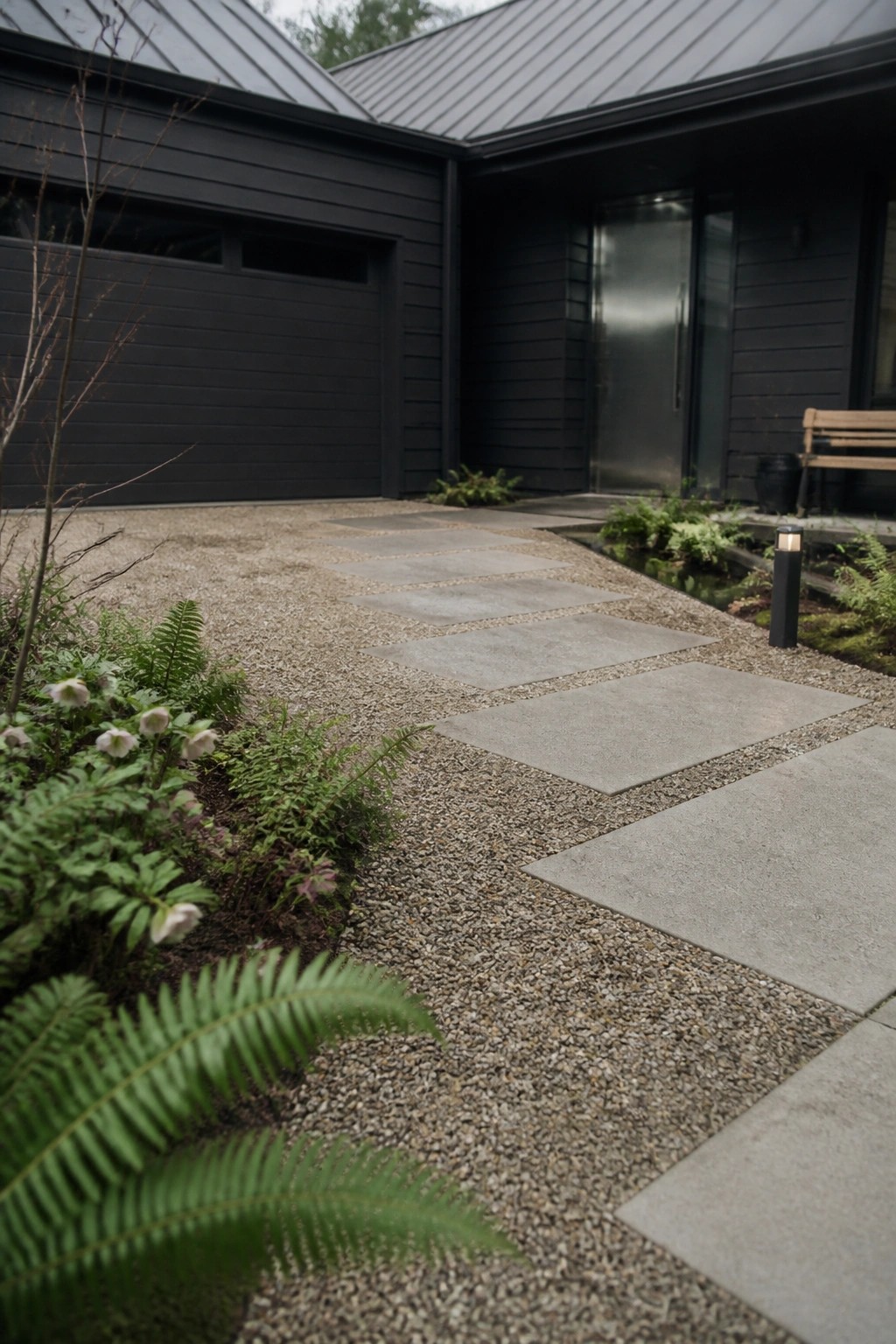 A dark modern house exterior with a pea gravel driveway lined by rectangular concrete pavers that lead toward the entrance, with ferns and other plants along the edges.