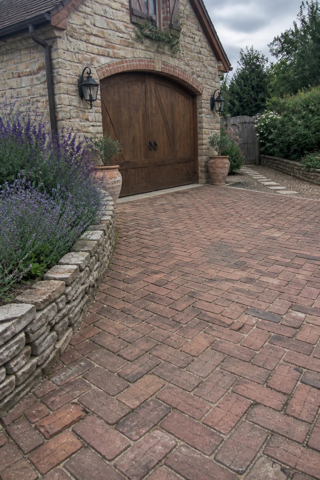 A red brick driveway in herringbone pattern leads to wooden garage doors on a stone house, bordered by a low stone wall and purple flowering plants.
