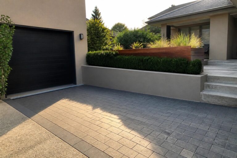 A gray interlocking paver driveway leading to a black garage door on a modern house with landscaping on the left.