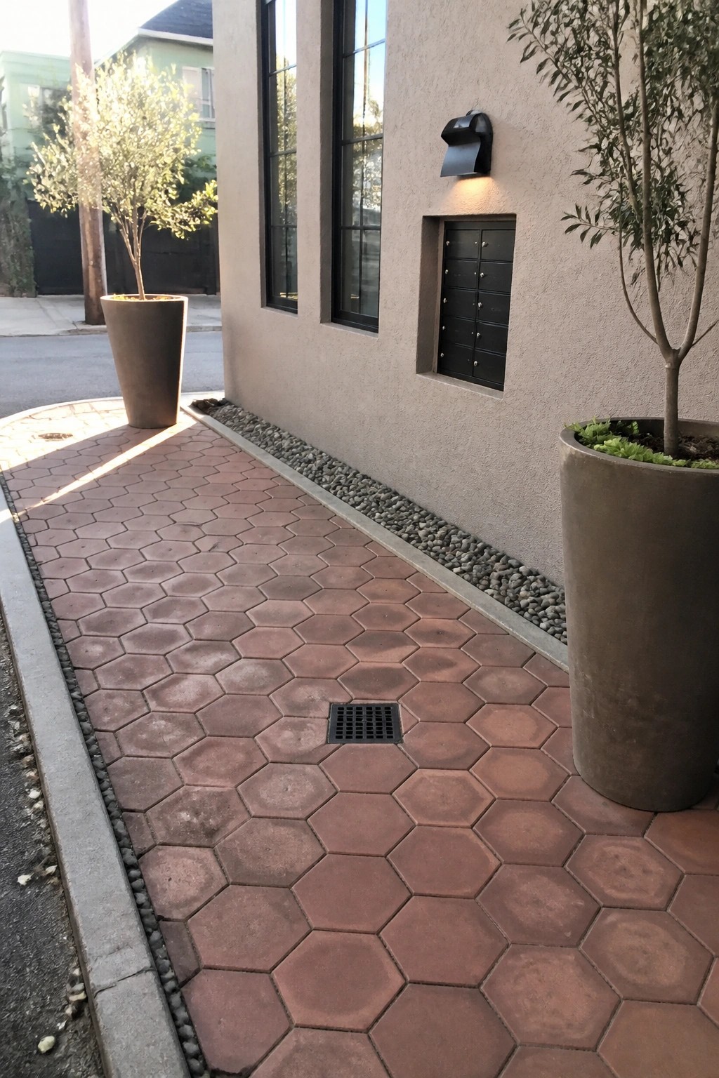 Hexagonal brick walkway beside stucco wall with potted trees and drain