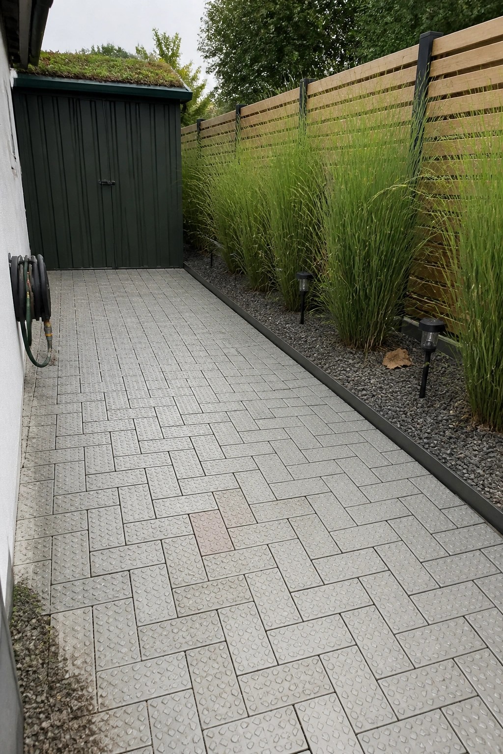 A narrow paved path of gray interlocking tiles in a herringbone pattern runs beside a dark green shed and a wooden fence lined with tall ornamental grass.