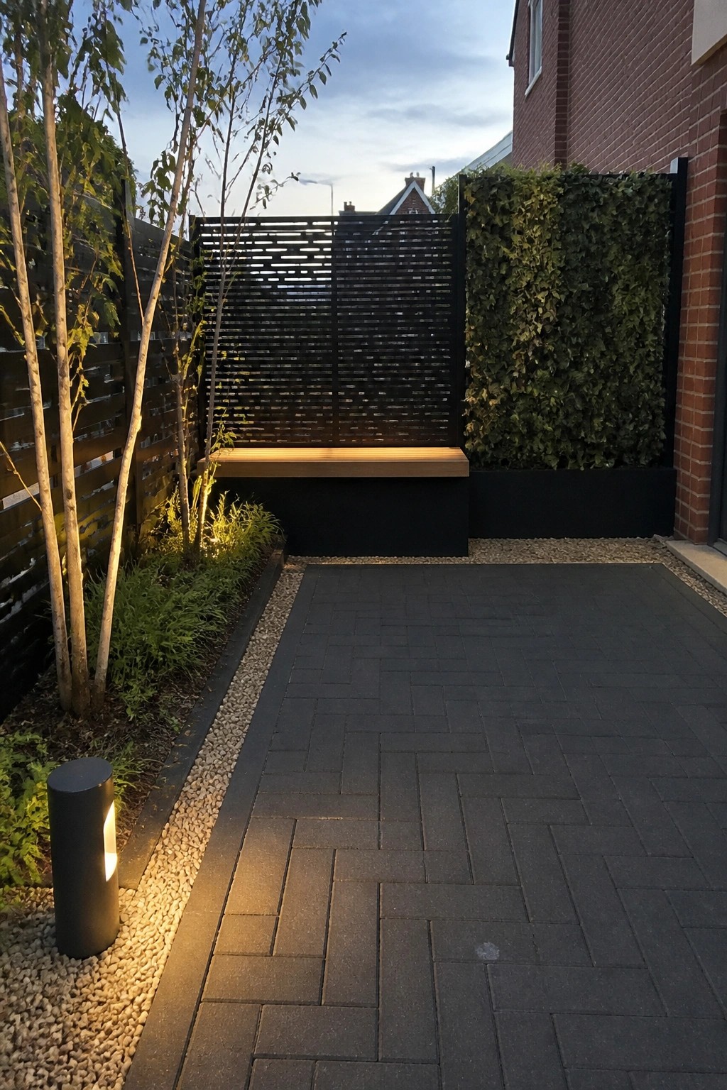 A paved outdoor courtyard with dark interlocking tiles, a built-in bench, gravel beds with young trees, and a vertical garden wall beside a brick wall.