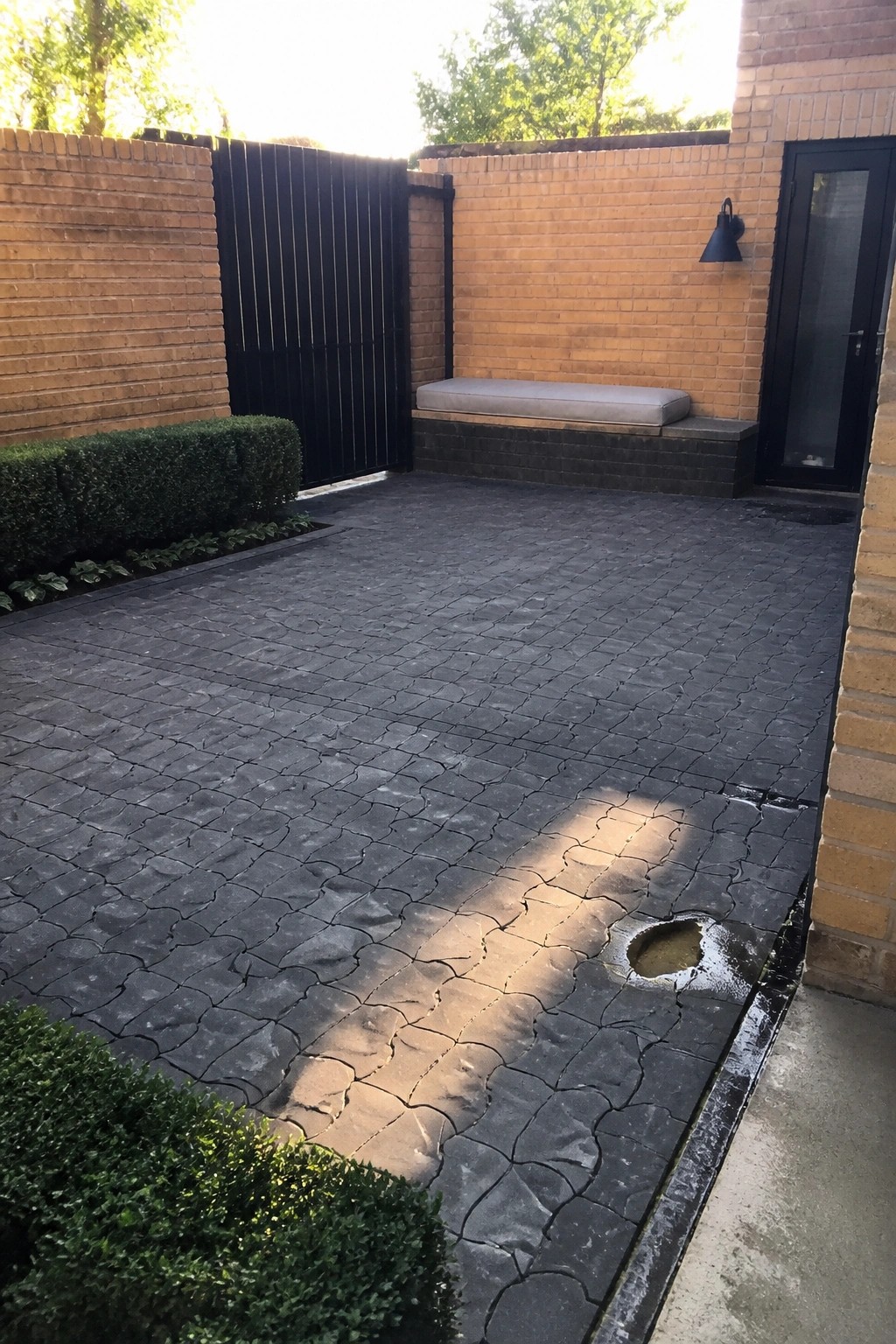 A dark gray interlocking tile courtyard bordered by brick walls, a black gate, and a built-in bench.