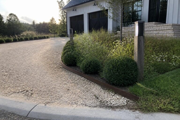 A gravel driveway with a curved metal edging separating it from planted beds containing shrubs and two wooden light posts.