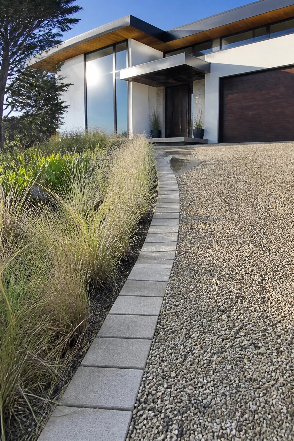 Modern house exterior with gravel driveway and a curved border of rectangular gray stone pavers separating it from garden beds with tall grasses.