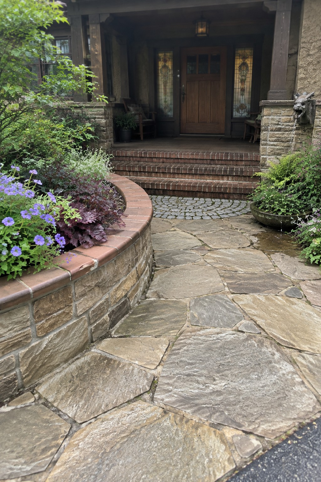 A curved stone pathway of irregular slabs leads to brick steps at a house entrance, bordered by a brick-edged garden bed with purple and green plants.