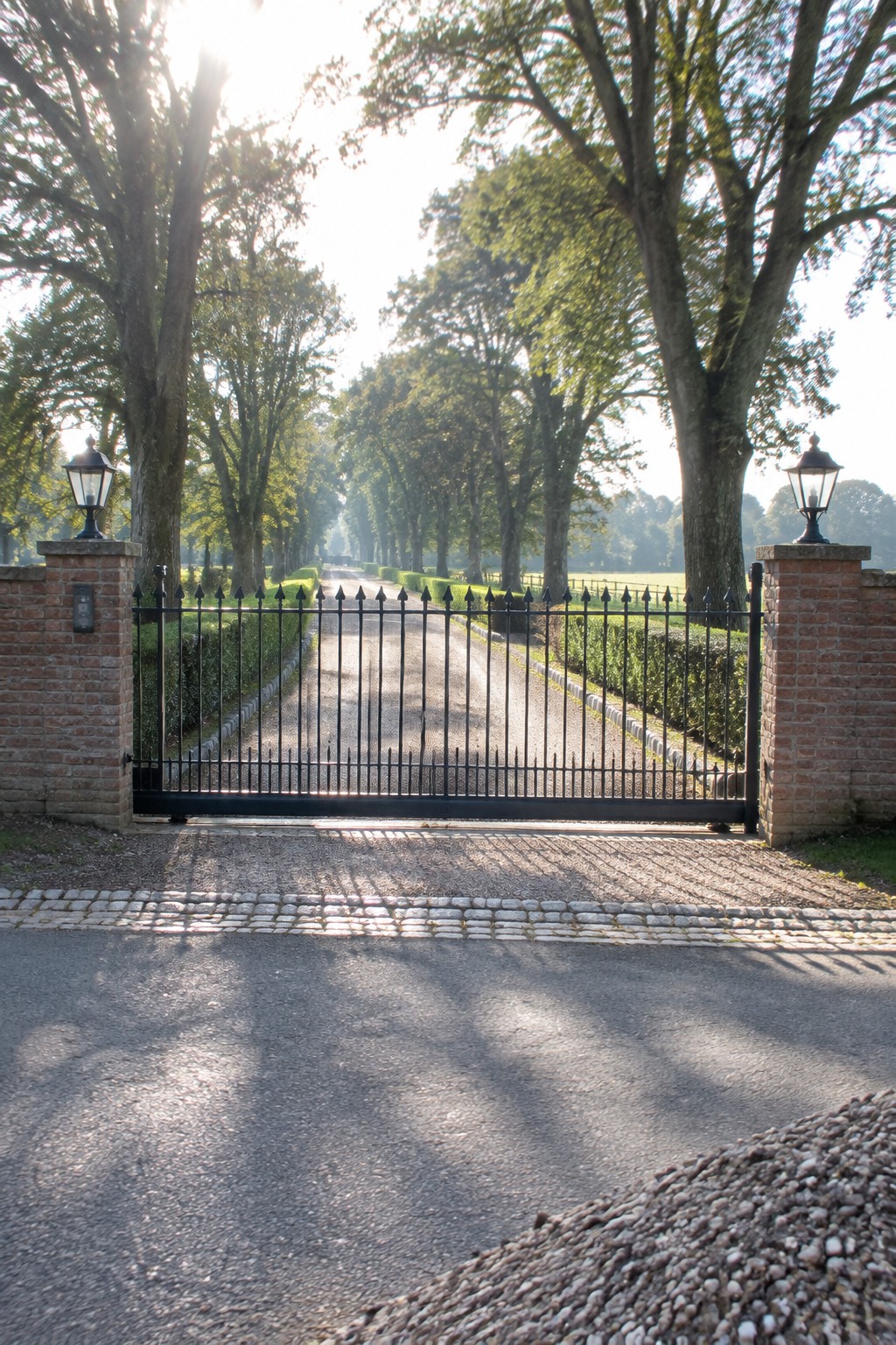 A black wrought iron driveway gate stands closed between two brick pillars with lanterns, opening onto a gravel road lined with tall trees.