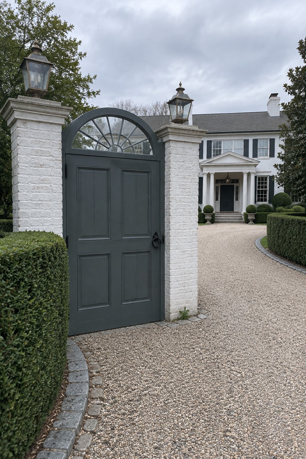 A dark gray arched double gate with a fanlight transom is set between white brick pillars at the entrance of a white colonial house with a gravel driveway.