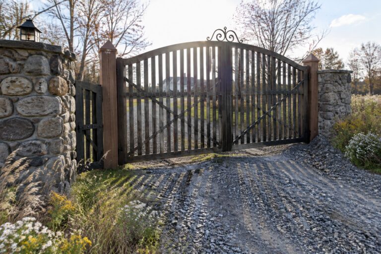 Open black metal gate between stone pillars on gravel drive amid wildflowers and trees