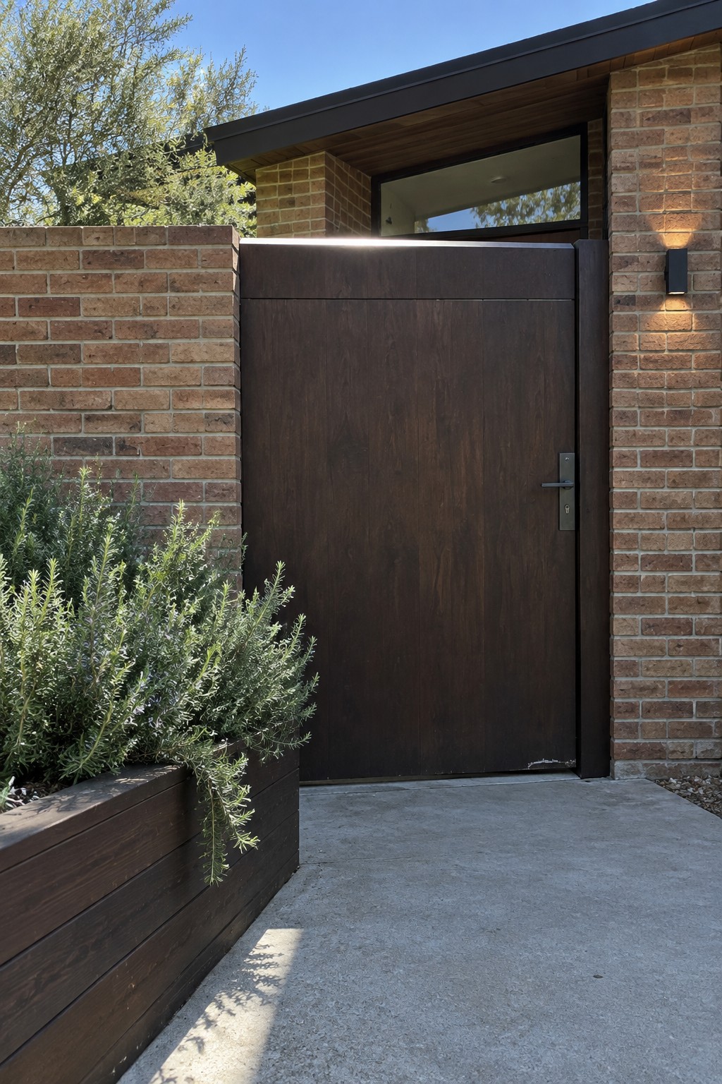 A dark wood single-panel driveway gate installed between brick walls next to a house, with a concrete driveway and a wooden planter box holding green shrubs in the foreground.
