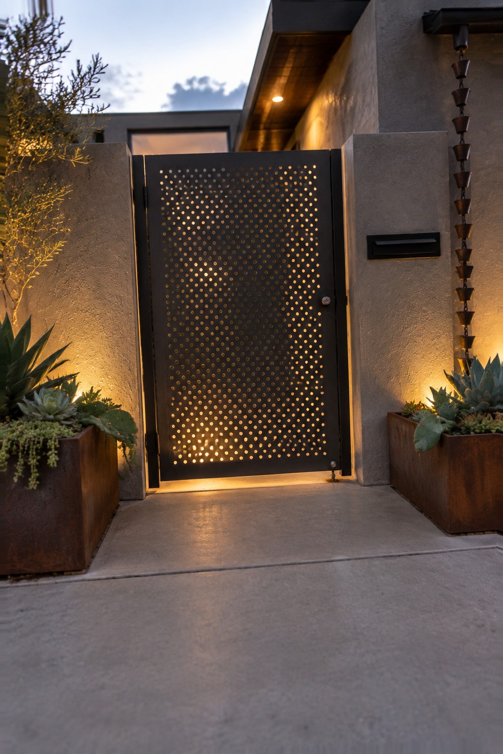 A black metal driveway gate with a pattern of small circular perforations stands between concrete walls, with succulent planters on both sides.
