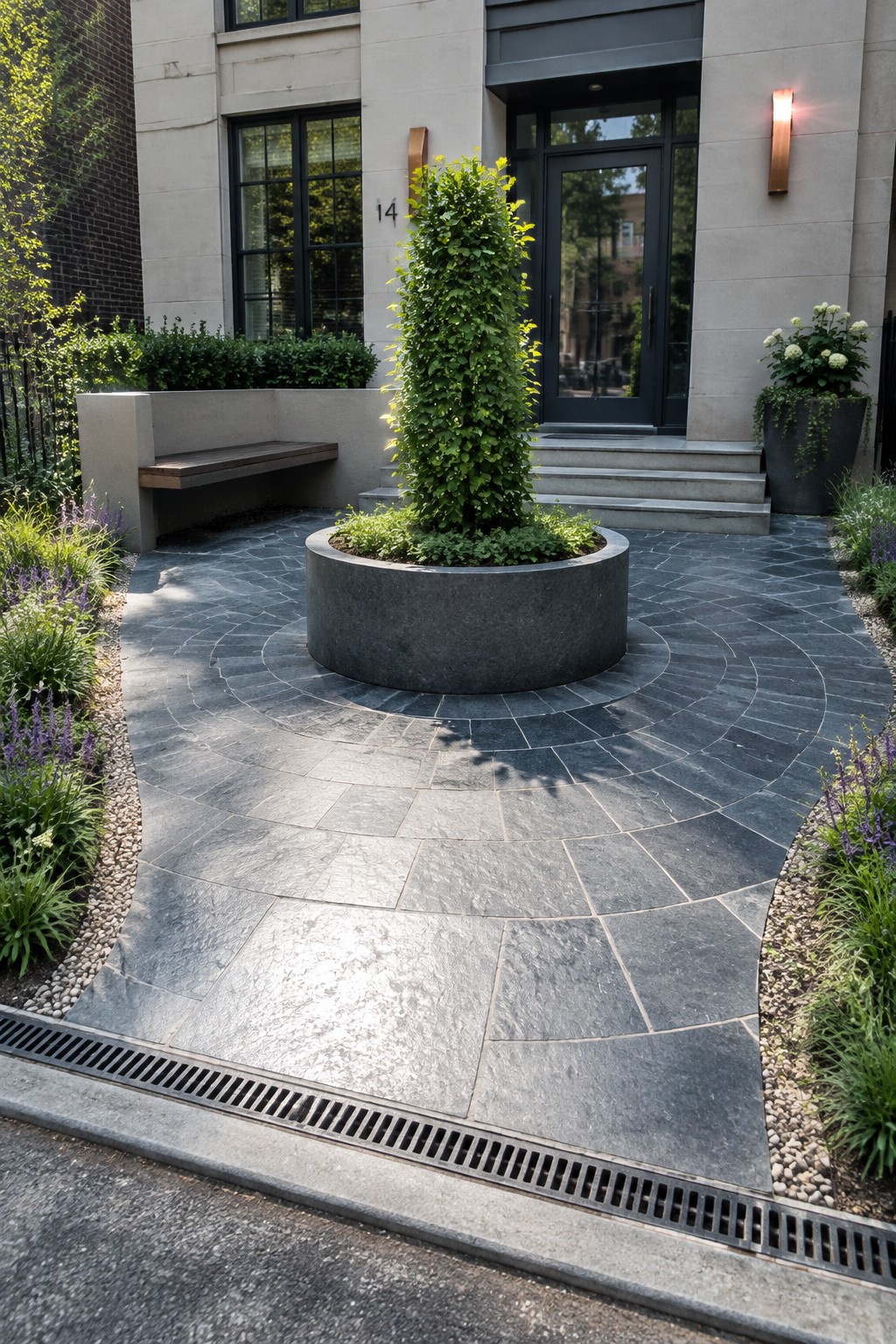 A modern house entrance with a circular driveway of gray stone pavers and a large central round planter holding a tall shrub, surrounded by low garden beds.