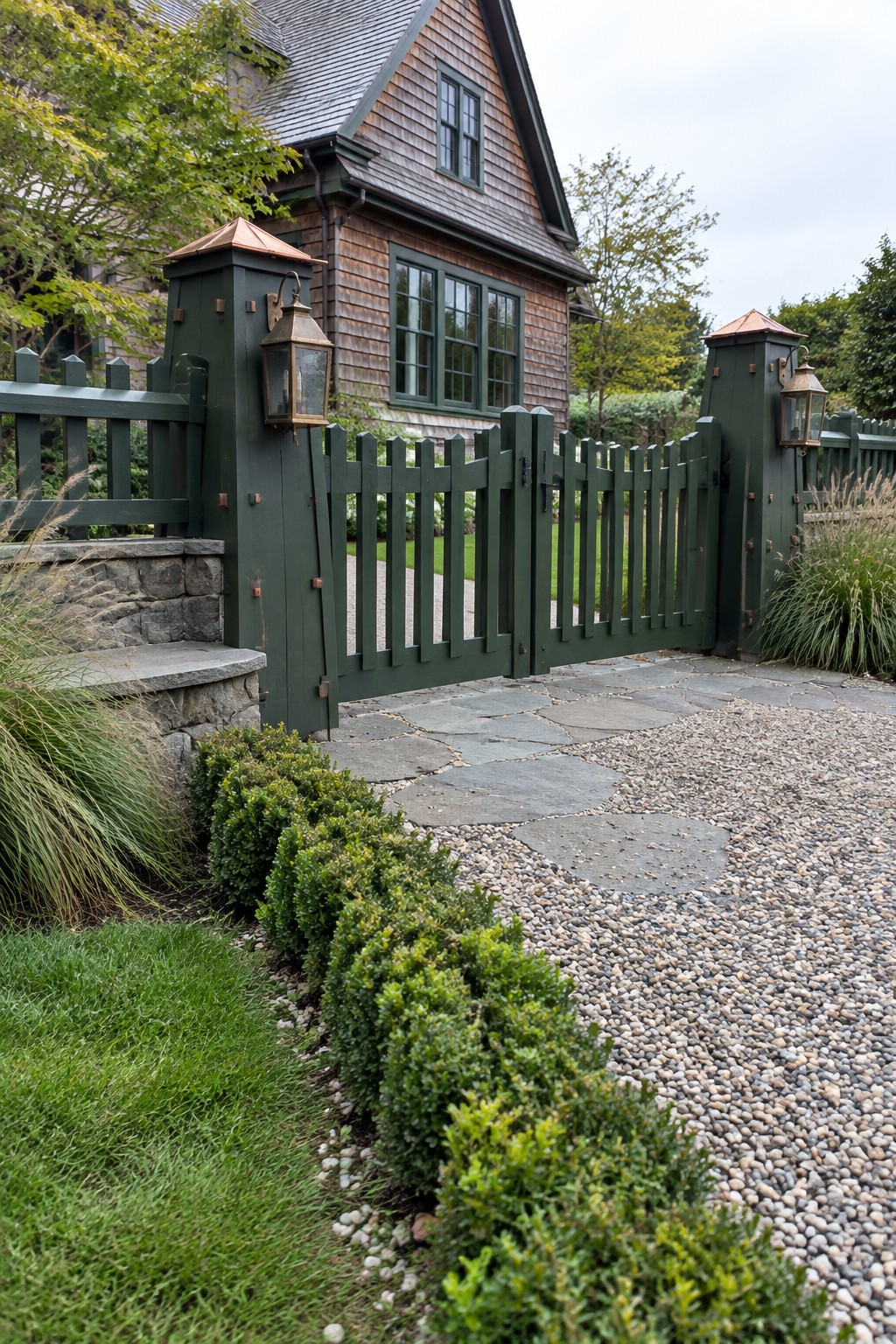 A dark green wooden driveway gate with copper-topped posts and lanterns stands open beside a shingled house with green trim.