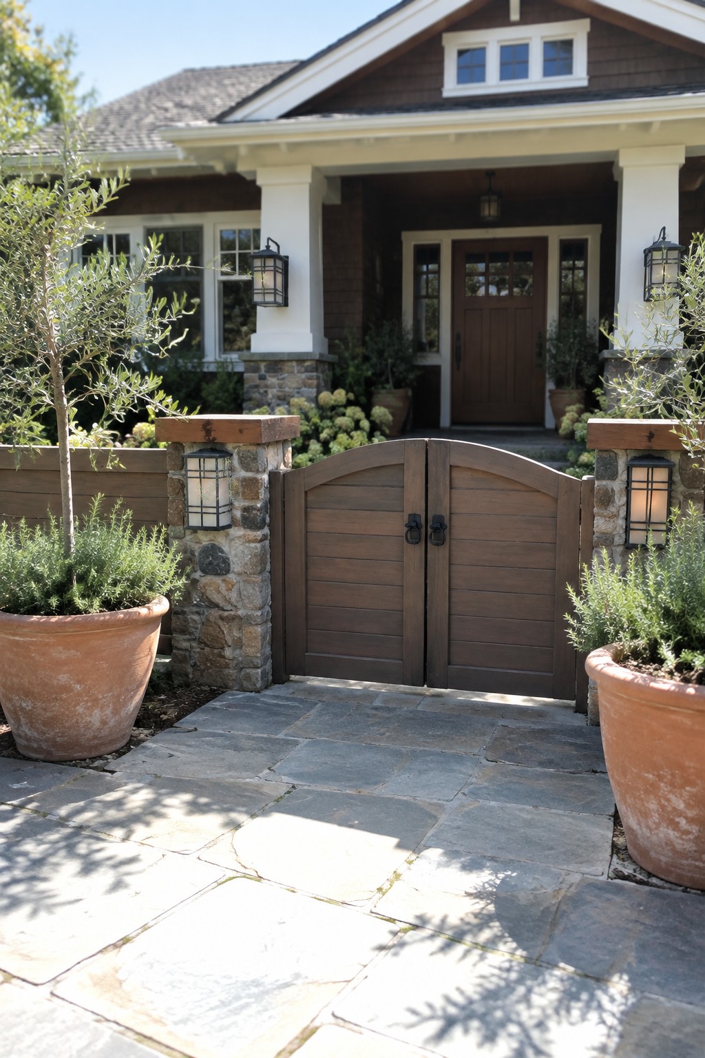 Wooden arched gate between stone pillars with lanterns and potted plants on walkway to house