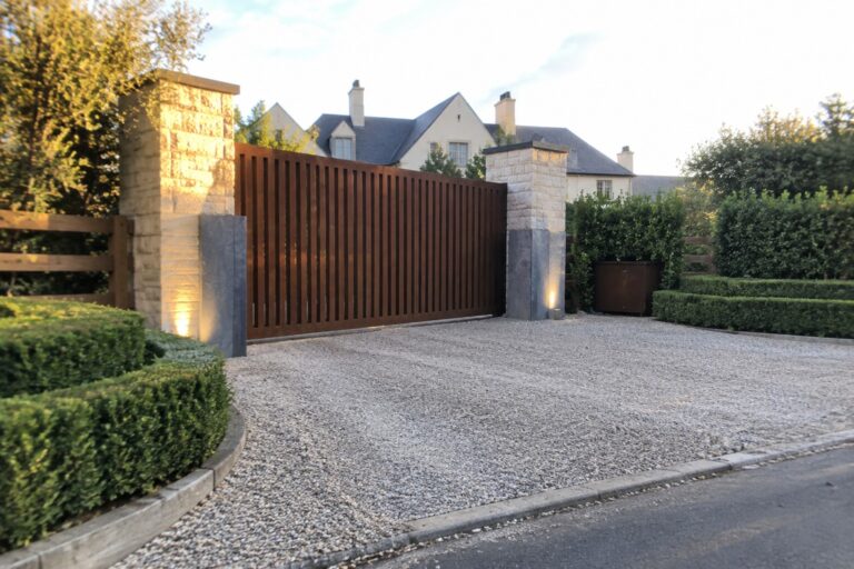 13 Grand Large Driveway Gate Ideas for Spacious Properties Brown slatted gate between illuminated stone pillars on gravel driveway