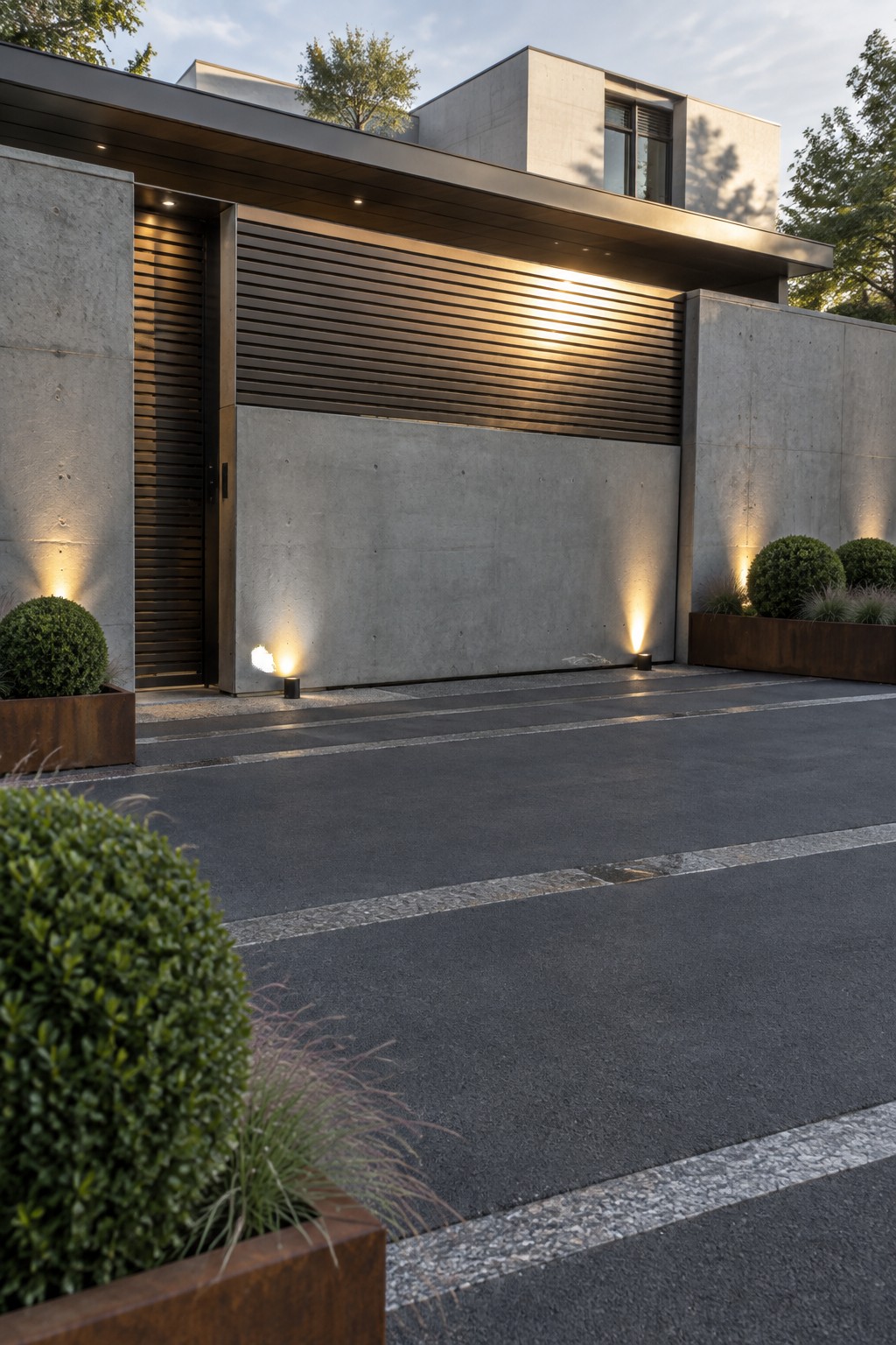 Modern concrete house with a wide horizontal slatted metal gate spanning the driveway, flanked by concrete walls and ground lighting at dusk.