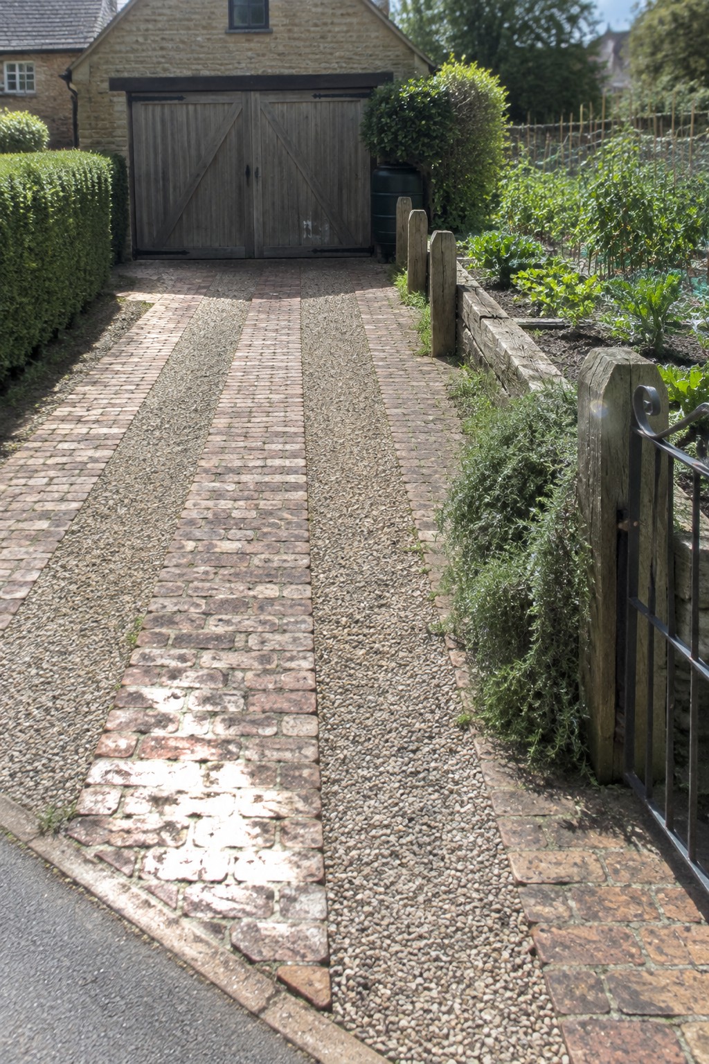 A gravel and brick driveway path leading to wooden garage doors on a stone cottage, with garden beds and a wooden gate visible on the right side.