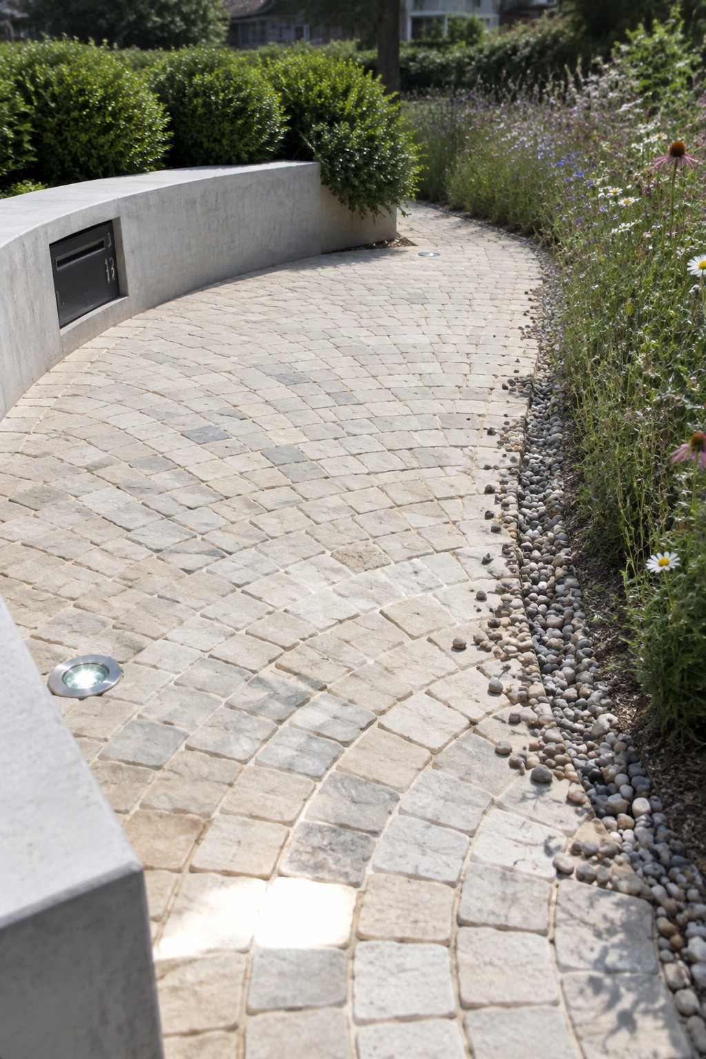 A curved driveway of light stone pavers with gravel edging on the right and a low concrete wall with a built-in mailbox on the left.