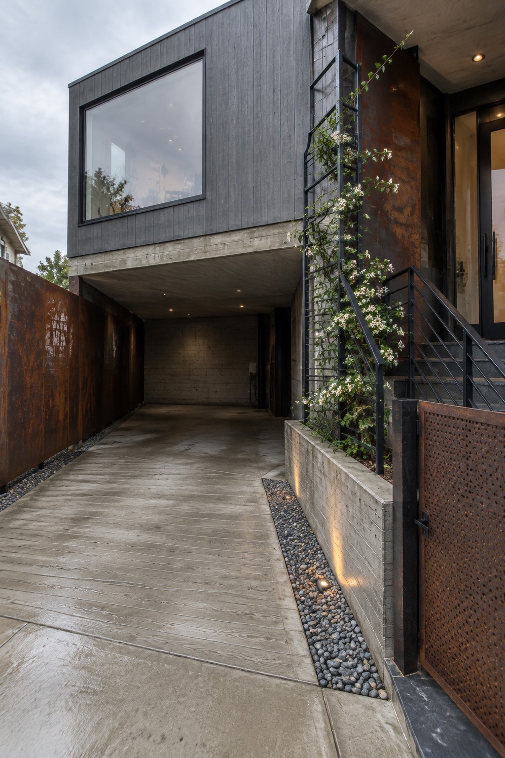 Modern house exterior showing a narrow concrete driveway bordered by rusted metal panels on the left and a low concrete planter with black metal railing on the right.