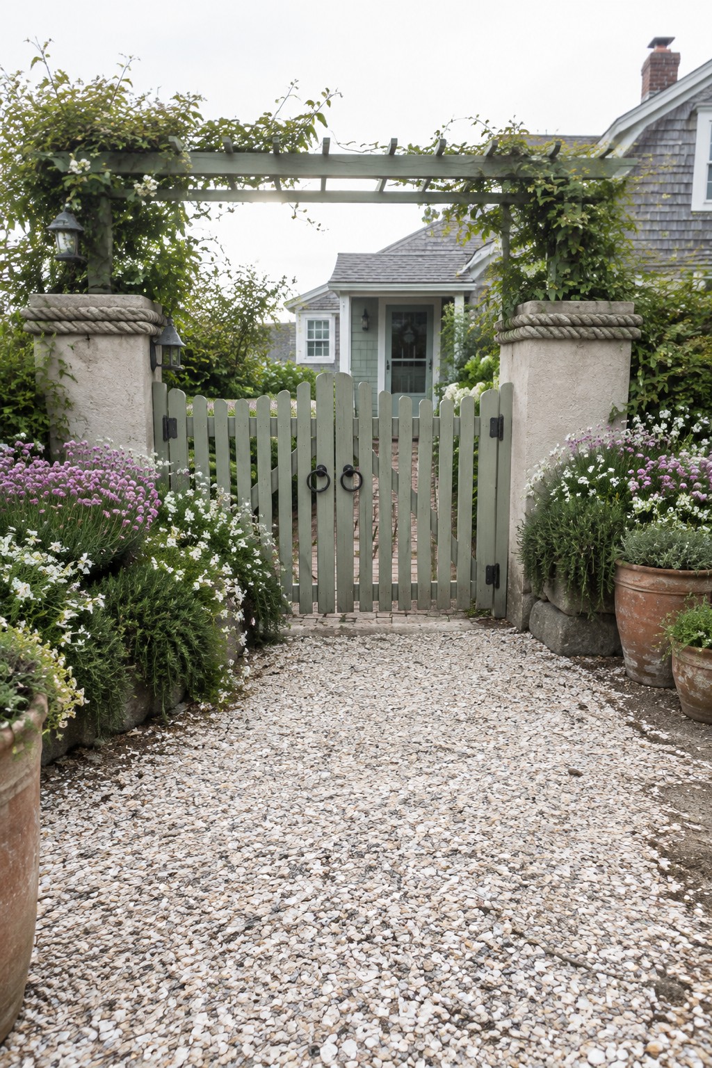 A green wooden gate with black hardware sits between two stone pillars under a pergola covered in green vines, with a gravel path leading toward a light-colored house.