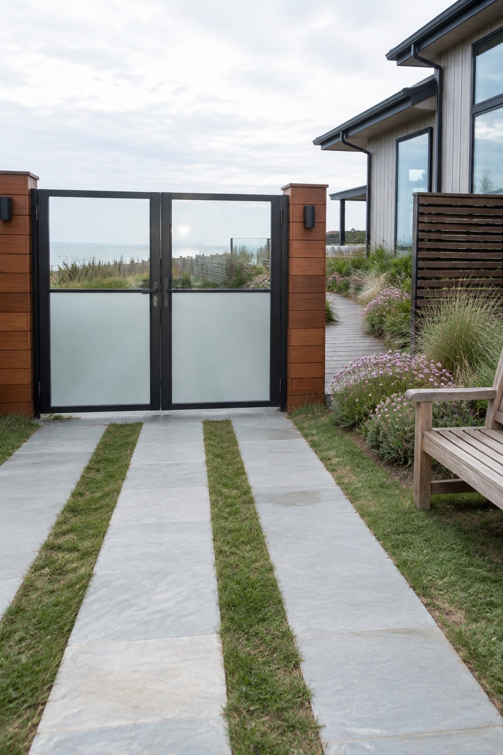 A modern double driveway gate with black metal frames, clear upper glass, and frosted lower panels stands between wooden posts at the entrance to a coastal home.