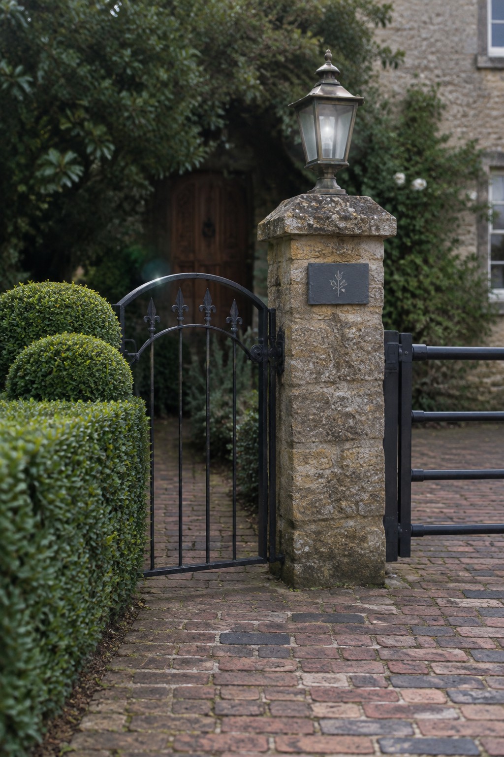 A black metal driveway gate with decorative tops stands open beside a weathered stone pillar with a lit lantern, next to a brick path and trimmed hedges.