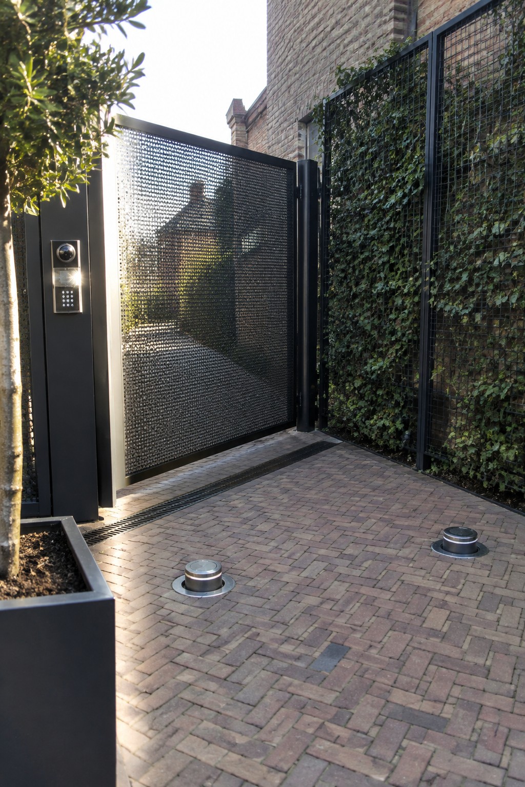A black perforated metal driveway gate stands partially open on a brick-paved surface beside an ivy-covered wall and a wall-mounted intercom panel.