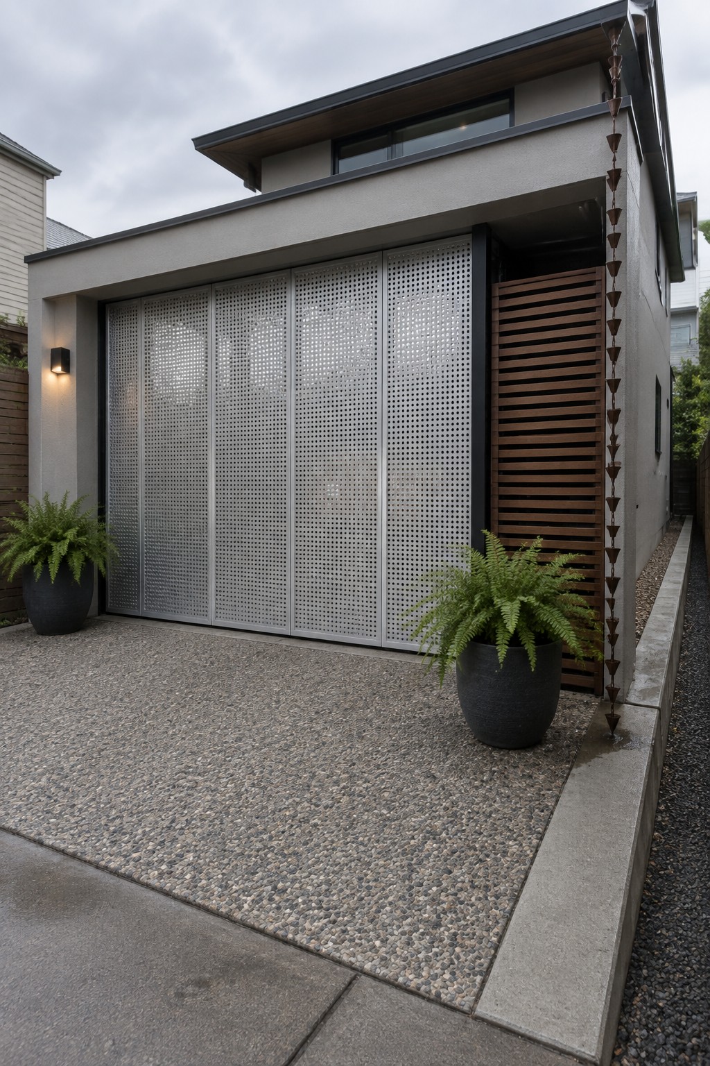 Modern house exterior with a wide driveway gate made of perforated metal panels next to a wooden slatted section, with potted ferns on gravel ground.
