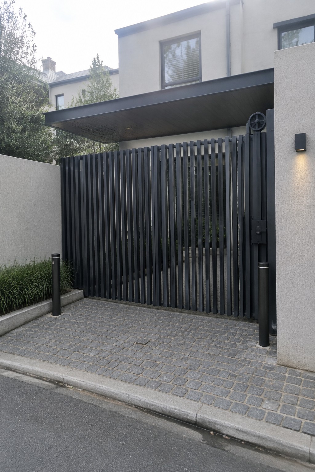 A black vertical slat driveway gate with an overhead canopy and two bollards on a cobblestone surface in front of a modern light-colored house.