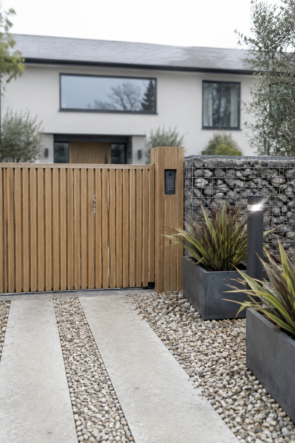 A modern wooden driveway gate with vertical slats stands beside a gabion wall, concrete planters, and a gravel path leading to a white house.