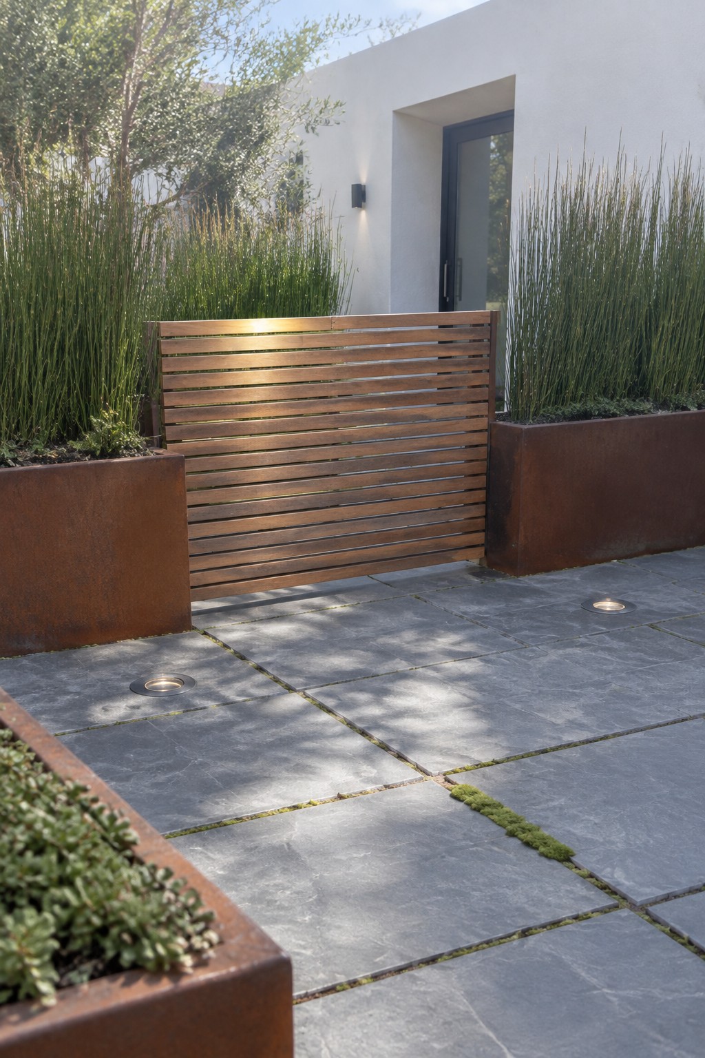 Modern patio with wooden slat screen, rusted planters, tall grasses, and stone pavers