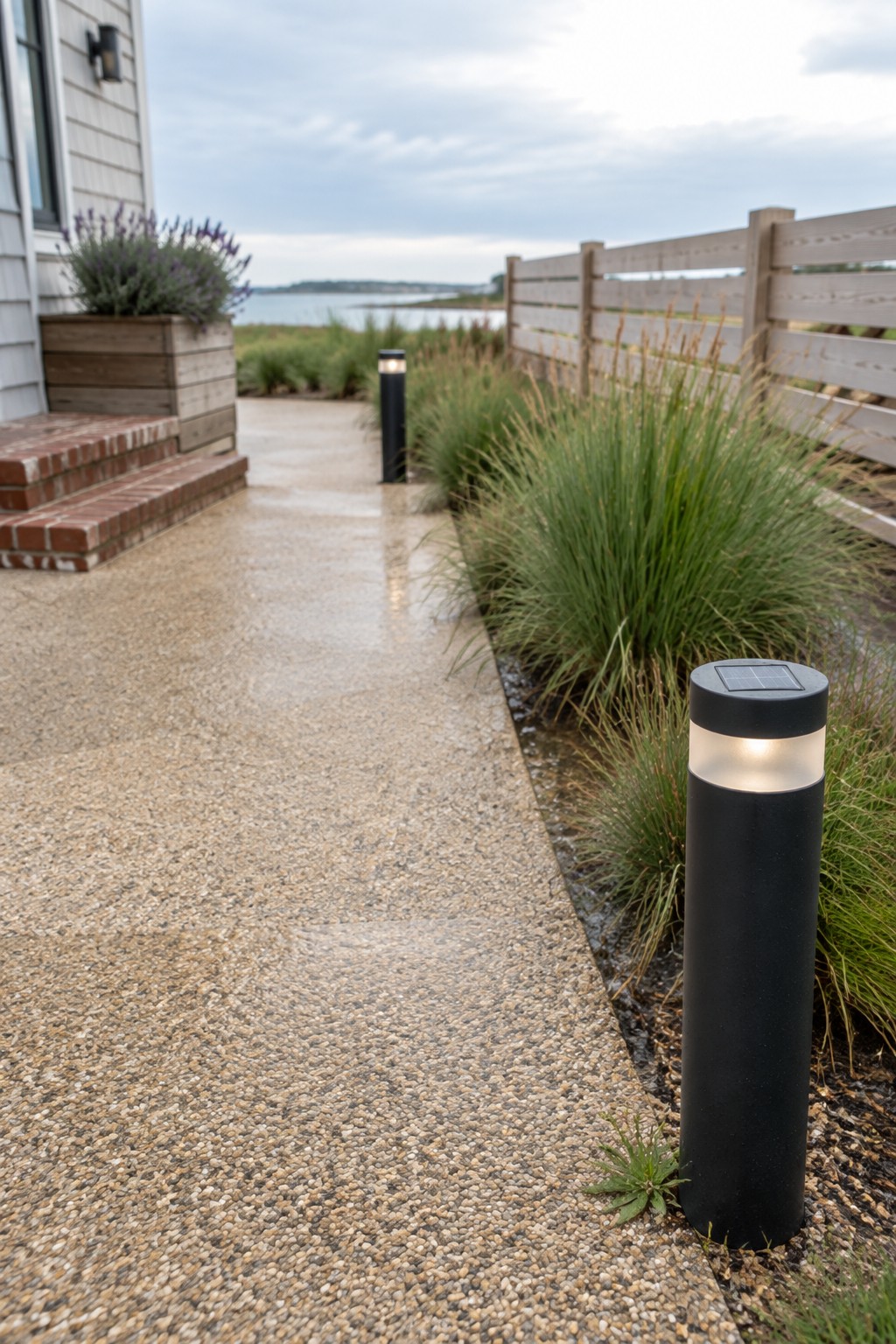 A pebbled pathway beside a house lined with black solar lights and tall grasses next to a wooden fence.
