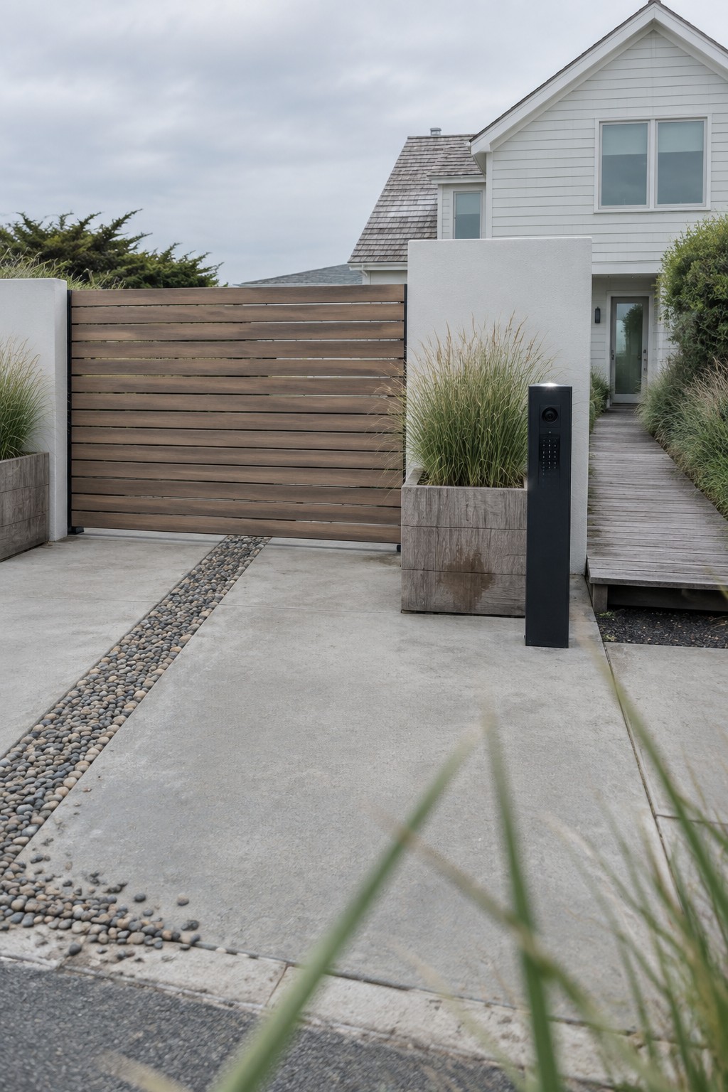 A modern white house exterior with a horizontal wooden slat driveway gate, concrete driveway with pebble strip, wooden walkway, and a black automated entry post with keypad and camera.