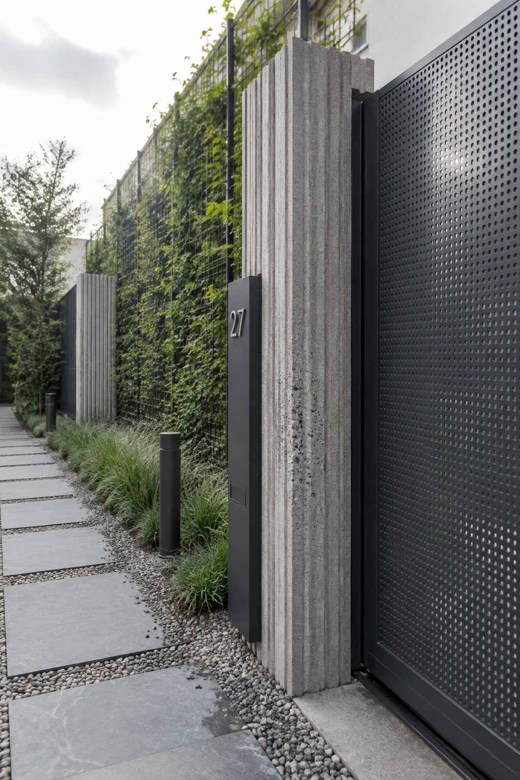 A modern black perforated metal driveway gate stands open beside a tall gray fluted concrete pillar with the number 27 mounted on a dark panel, next to a stone path and green living wall.