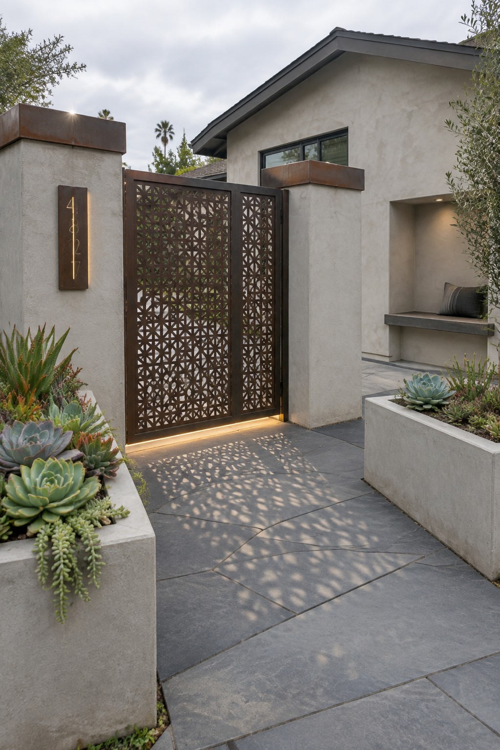A modern home entrance with a brown steel driveway gate featuring a geometric cutout pattern, flanked by concrete pillars, stone pavers, and succulent plantings.